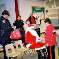 12/15/06 - Christmas at Mission Educational Center, Police Officer “Nacho” Martinez as Santa - Santa greeting students and handing them gifts as his Policer Officer escorts, and others, look on.