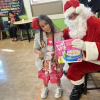 12-18-24 - 43rd Annual Lions Christmas with Santa at the Mission Education Center with Los Bomberos Firefighters - One by one, the students receive their presents from Santa, each having a quick pose after with Santa and his helper. Lion Zenaida Lawhon sits in the background.