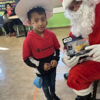 12-18-24 - 43rd Annual Lions Christmas with Santa at the Mission Education Center with Los Bomberos Firefighters - One by one, the students receive their presents from Santa, each having a quick pose after with Santa and his helper. Lion Zenaida Lawhon sits in the background.